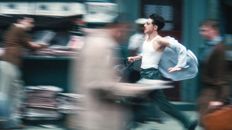 Eine dynamische Unschärfeaufnahme eines Mannes im weißen Unterhemd, der mit einer Tasche durch eine belebte Stadtstraße rennt.