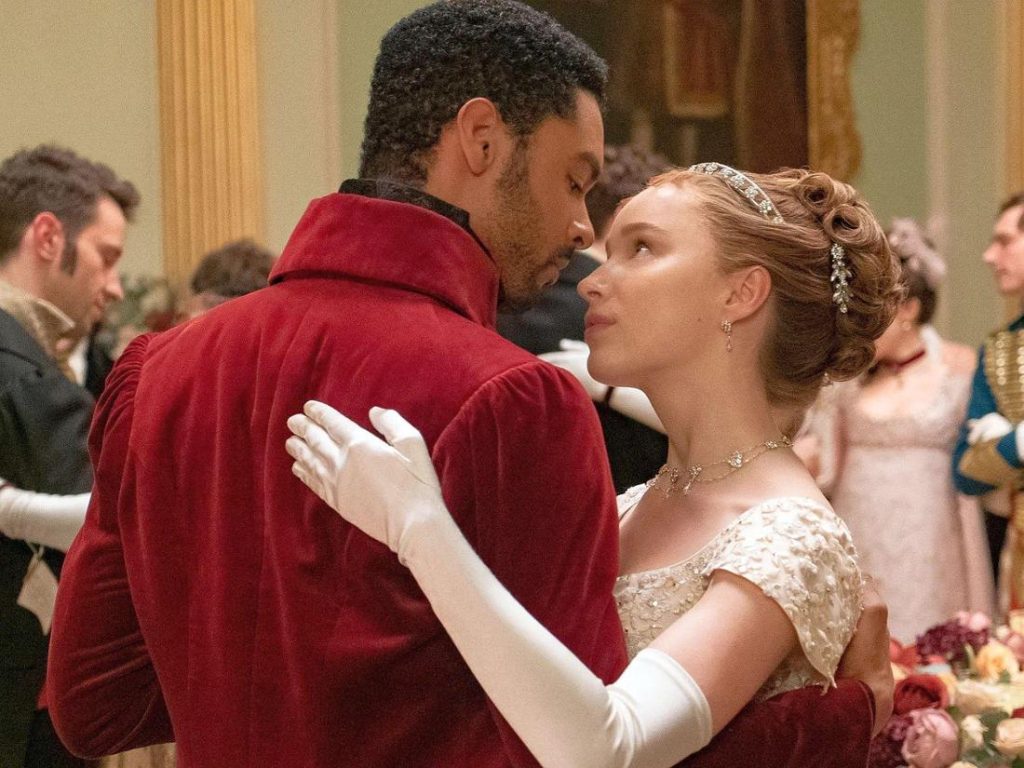 Auf einem Ball tanzen ein Mann im roten Frack und eine Frau im weißen Kleid. Sie blickt zu ihm auf, ihre Arme um ihn gelegt. Andere Ballgäste im Hintergrund.