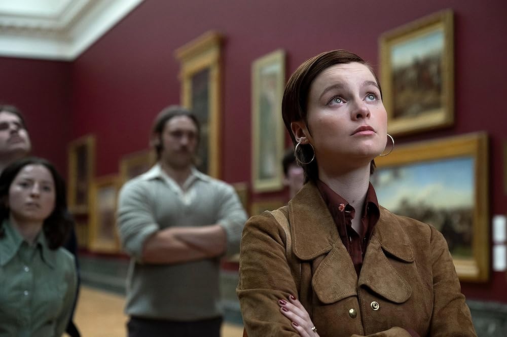 Junge Frau mit brauner Wildlederjacke und Creolen blickt in einem Museum nach oben. Rote Wände mit Gemälden. Unscharfe Personen im Hintergrund.