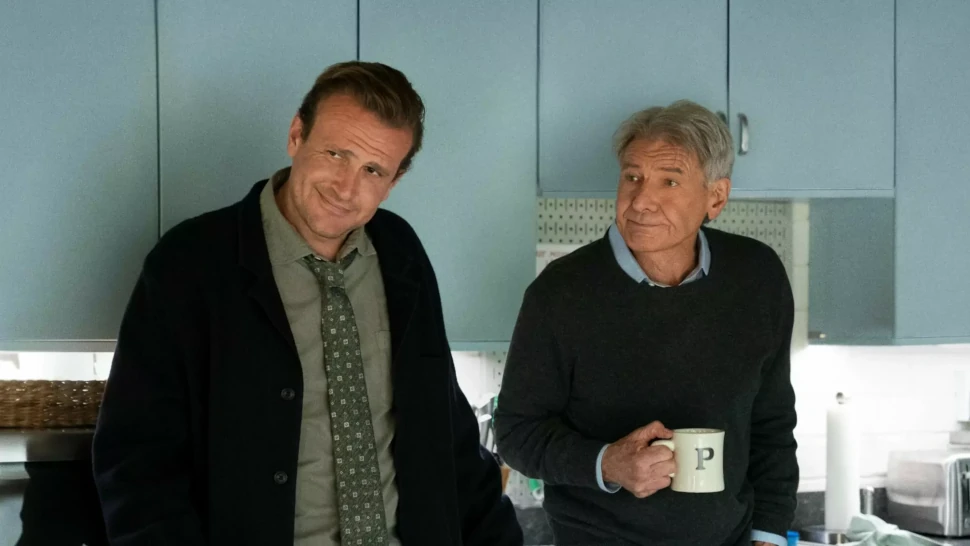 Zwei Männer stehen in einer hellblauen Küche; einer lächelt verschmitzt, der andere hält eine Tasse.