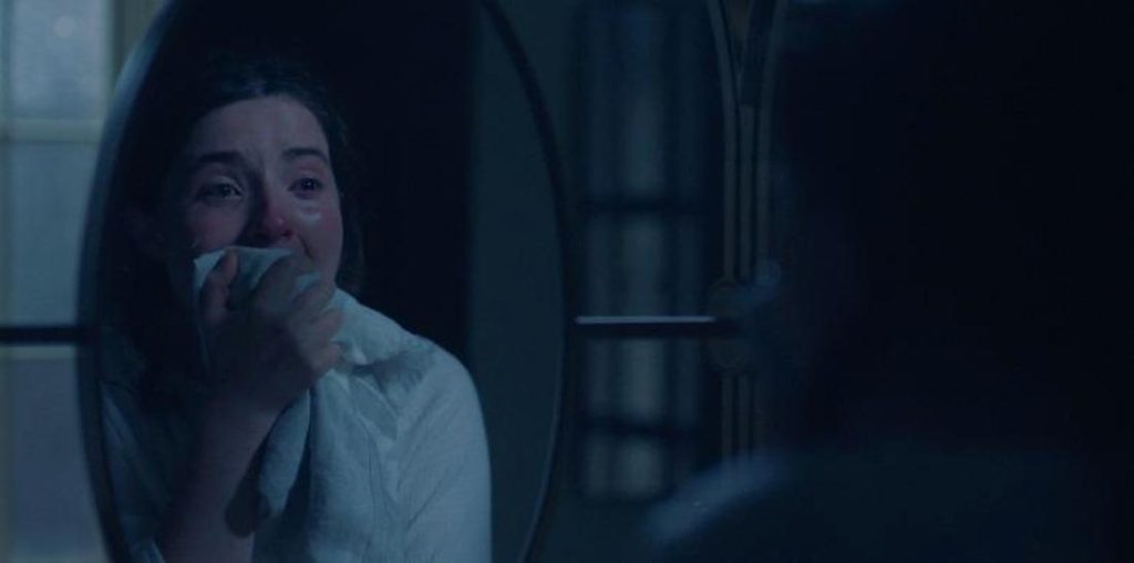 Im Spiegel weint eine Frau in ein weißes Tuch. Ihr gerötetes, tränennasses Gesicht ist im dunklen Raum sichtbar, rechts eine Kopfsilhouette.