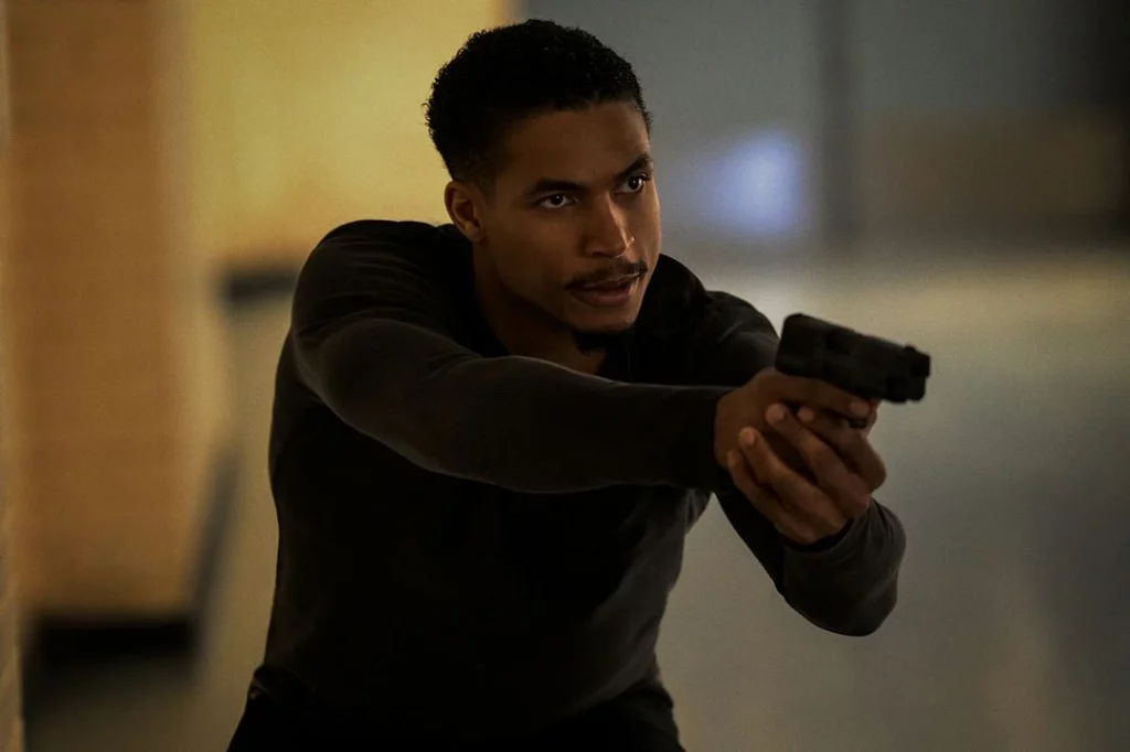 Ein Mann in dunkler Kleidung zielt konzentriert mit einer Pistole in einem schwach beleuchteten, gelblichen Flur.