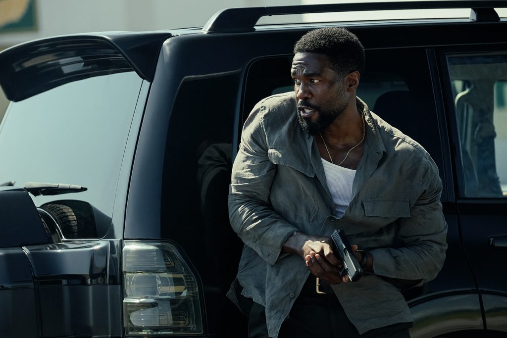 Ein bärtiger Mann steigt mit einer Pistole im Anschlag und wachsamem Blick aus einem schwarzen Geländewagen.