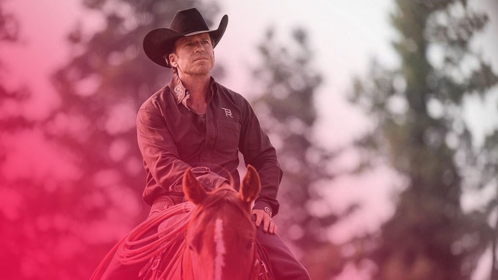 Ein Cowboy mit schwarzem Hut und braunem Hemd reitet auf einem Fuchs-Pferd. Er blickt konzentriert in die Ferne.