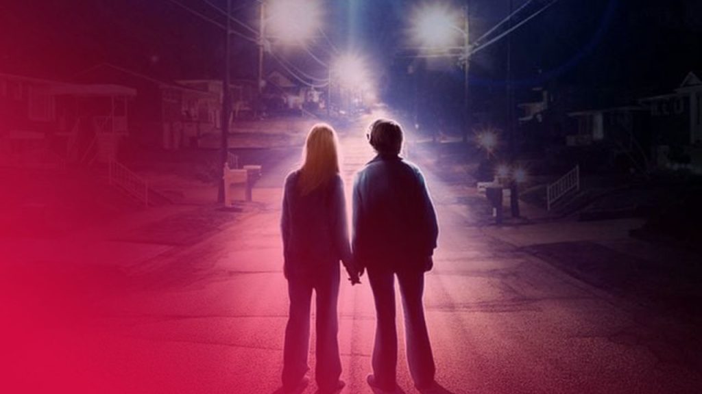 Rückansicht zweier Silhouetten, die sich an den Händen halten und in ein gleißendes Licht am Ende einer nächtlichen Straße gehen.