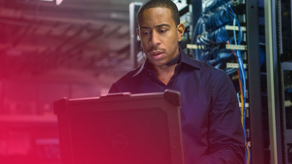 Ein Mann blickt konzentriert auf einen Laptop vor Server-Racks mit blauem Kabelsalat in einem technisierten Raum.