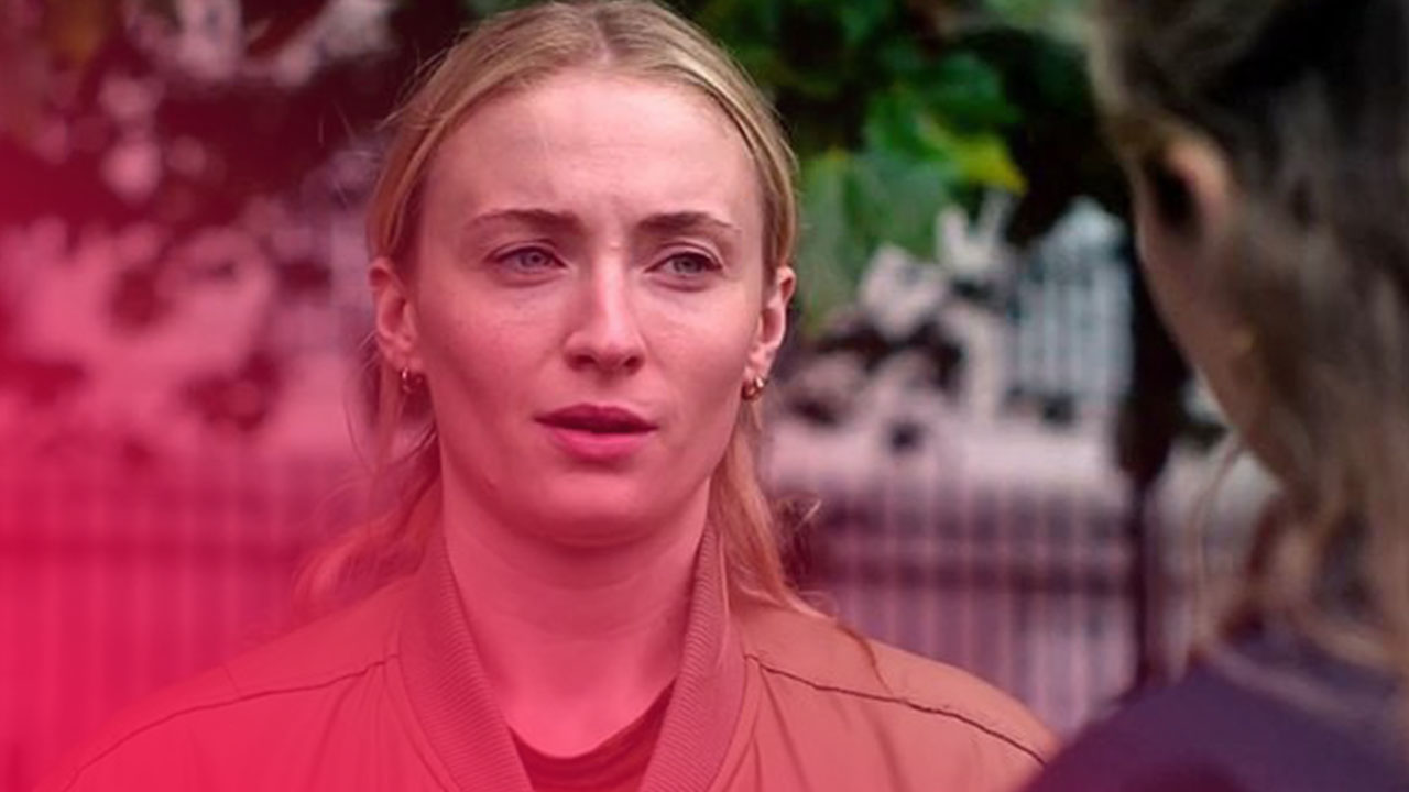 Nahaufnahme einer blonden Frau in einer beigen Bomberjacke, die mit besorgtem Blick zu einer Person außerhalb des Bildes spricht.