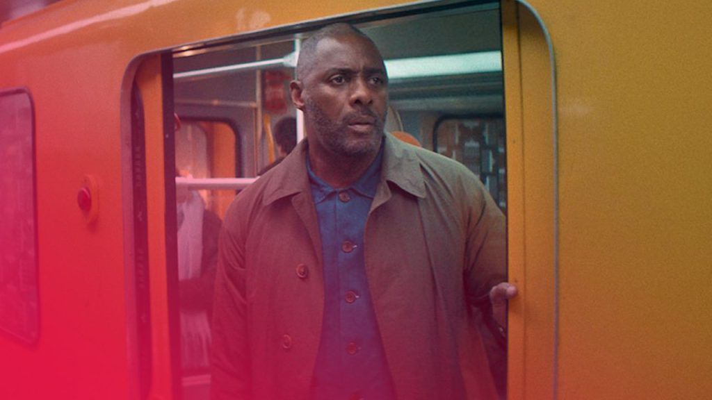 Ein Mann mit Bart, bekleidet mit einem blauen Hemd und einem olivgrünen Trenchcoat, steht in der offenen Tür eines U-Bahn- oder Zugwagons, der außen leuchtend gelb ist. Er blickt mit ernstem Ausdruck nach rechts aus dem Bild. Im Hintergrund des Wagens sind undeutlich weitere Personen und die Inneneinrichtung zu erkennen.