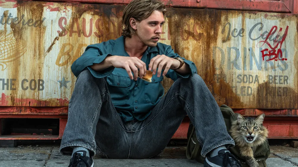 Ein Mann sitzt mit einem Snack vor einem rostigen Imbisswagen; neben ihm schaut eine flauschige Katze aus einer Tasche hervor.