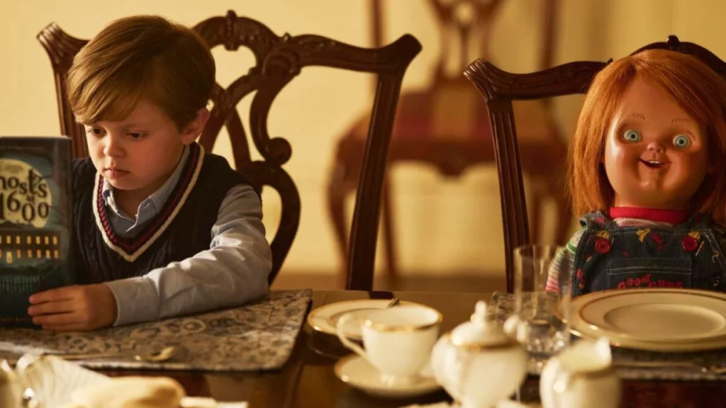 Ein kleiner Junge liest am Esstisch ein Buch, während rechts daneben die Chucky-Puppe auf einem Stuhl sitzt.