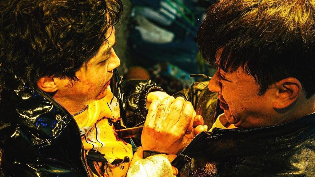 Zwei Männer im Nahkampf: Einer hält ein Messer, der andere greift dessen Hände. Beide wirken erschöpft und blutverschmiert.