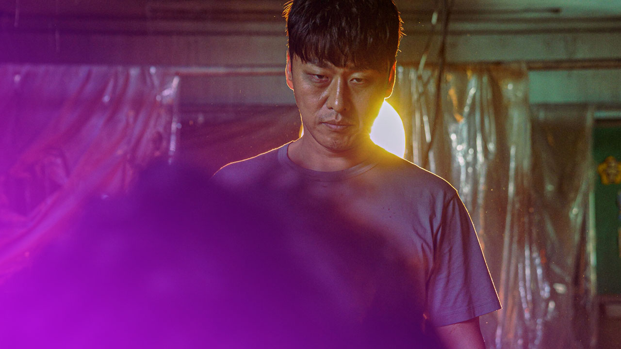 Ein Mann mit finsterem Blick steht in einem düsteren Raum; hinter ihm leuchtet eine helle Lichtquelle dramatisch auf.