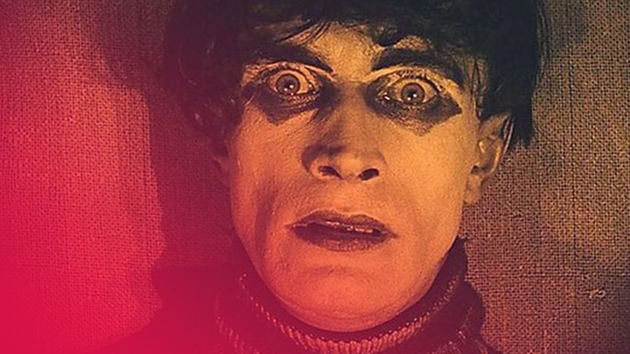 Nahaufnahme eines blassen Gesichts mit weit aufgerissenen Augen und dunklem Make-up vor grobem Stoffhintergrund.