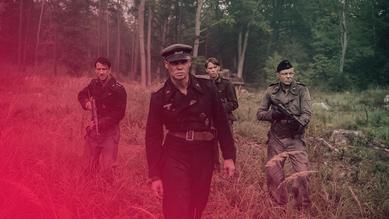 Vier erschöpfte Soldaten in dunklen Uniformen mit Gewehren gehen durch ein hohes Grasfeld vor einer Waldkulisse.