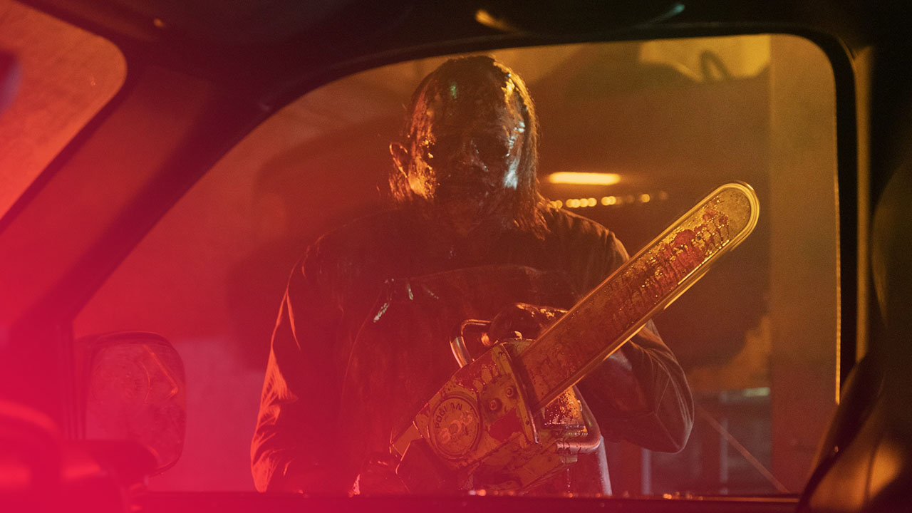 Eine furchterregende Figur mit einer blutigen Gesichtsmaske, die wie aus menschlicher Haut gemacht zu sein scheint, steht in einem leicht abgedunkelten Raum vor einem Auto. Die Figur trägt eine blutbefleckte Schürze und hält eine große Kettensäge, deren Klinge ebenfalls mit Blut bespritzt ist. Sie blickt direkt auf den Betrachter. Das Licht im Hintergrund ist gelblich-orange und schafft eine unheimliche Atmosphäre. Die Szene wird aus dem Inneren des Autos betrachtet, wobei der Seitenspiegel und die Windschutzscheibe zu sehen sind.