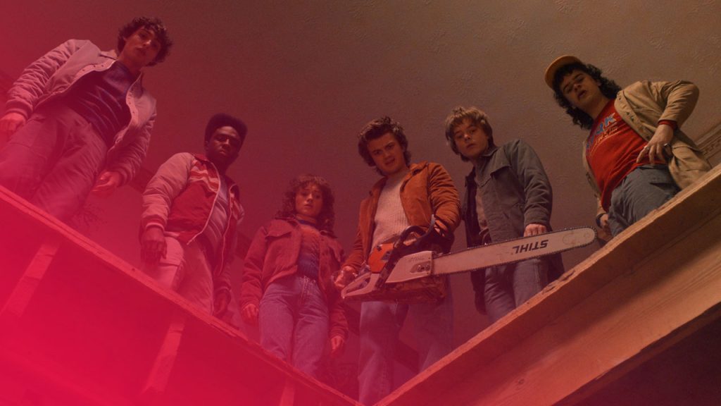 Sechs Personen im 80er-Jahre-Stil stehen auf einem Dachbalken und schauen nach unten. Eine Person hält eine Kettensäge. Die Perspektive ist von unten nach oben gerichtet.