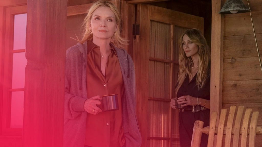 Zwei Frauen stehen vor einer Holzhütte; die vordere trägt eine braune Bluse und hält eine dunkle Tasse in der Hand.