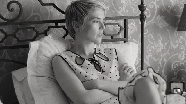 Eine Frau mit Pixie-Cut im gepunkteten Top lehnt nachdenklich in einem Bett mit gemusterter Tapete im Hintergrund.