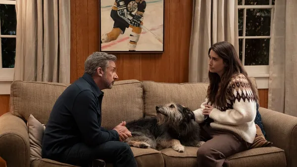 Mann und Frau sitzen auf einem Sofa und schauen sich an; ein struppiger Hund liegt entspannt zwischen ihnen.