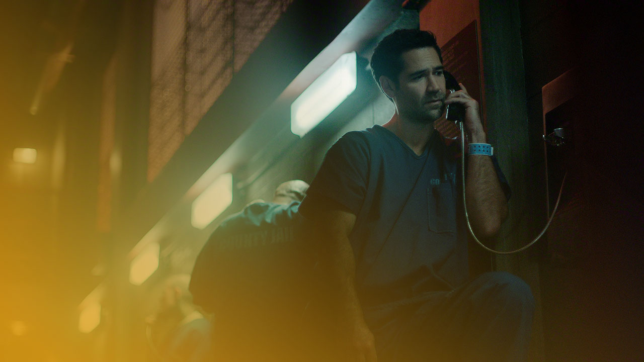 Mann in blauer Anstaltskleidung telefoniert besorgt in einem düsteren Gefängnisflur; kühles, grünes Licht.