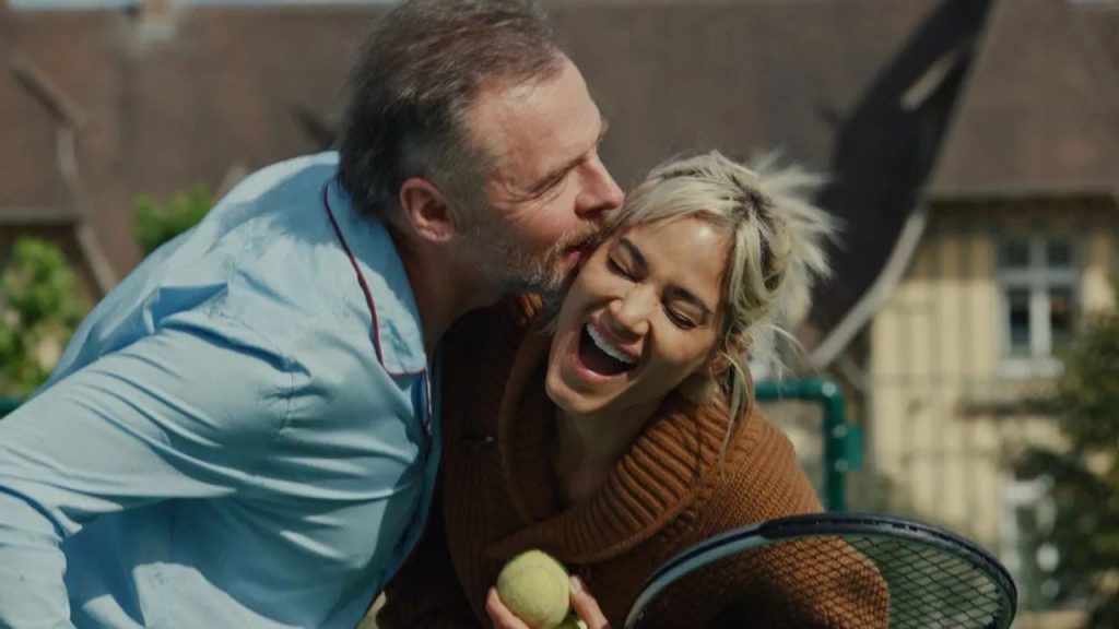 Simon Pegg küsst Sofia Boutella lachend auf die Wange, während sie einen Tennisball und einen Schläger hält.