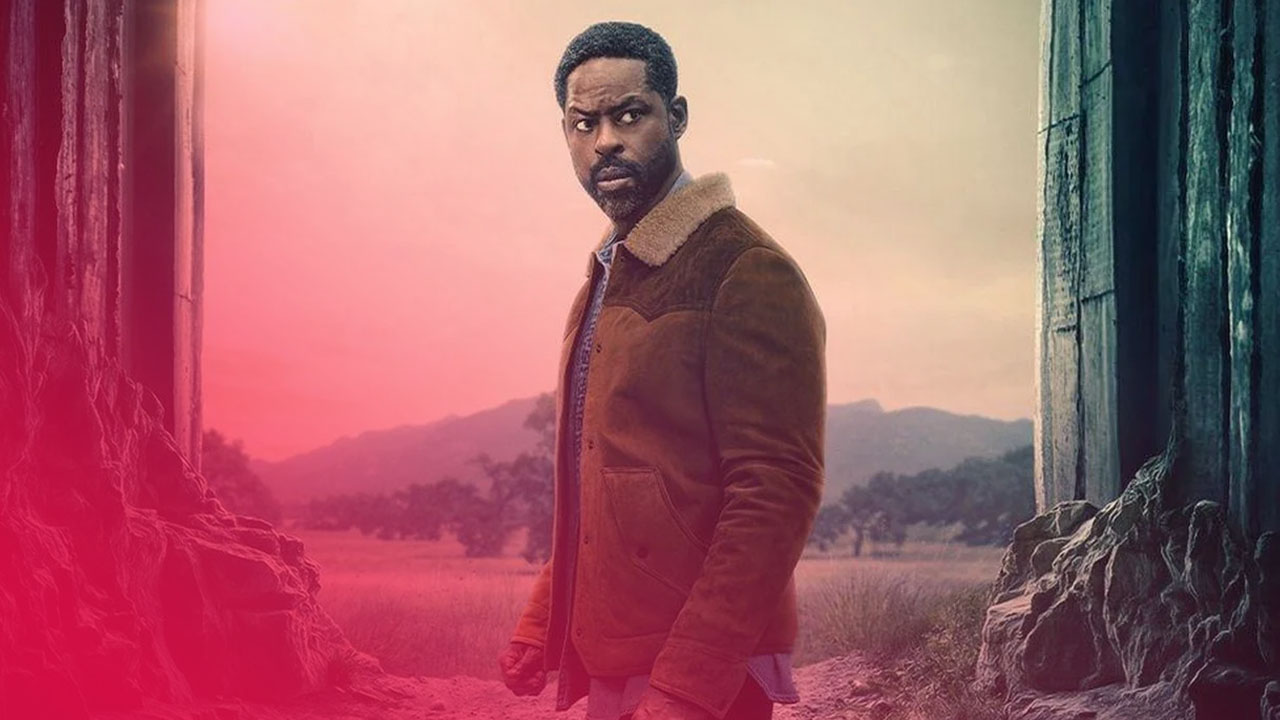 Ein bärtiger Mann im braunen Parka blickt ernst zwischen Felsen auf eine weite, hügelige Landschaft bei dämmrigem Licht.