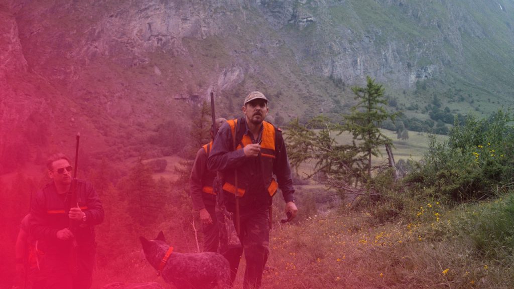 Jäger mit Gewehren und Jagdhund wandern an einem steilen, grünen Grashang vor einer massiven Felswand im Gebirge.