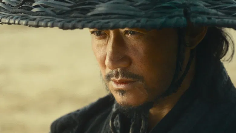 Porträt eines bärtigen Mannes mit dunklem geflochtenem Hut, der ernst nach unten blickt. Warmer, sandiger Hintergrund.