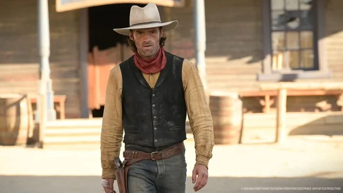 Ein Cowboy mit hellem Hut, schwarzer Weste und Revolvergurt geht vor einer Western-Kulisse mit Saloon.