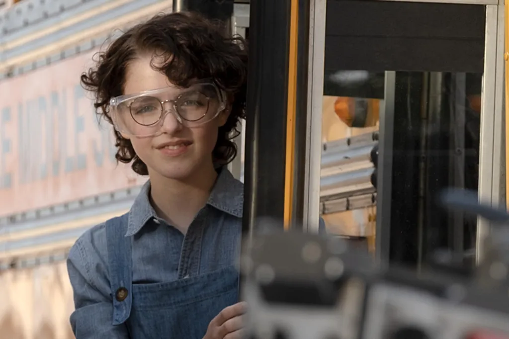 Lächelndes Mädchen mit lockigem Haar, Brille und Schutzbrille in einer Blaumann-Latzhose vor einem gelben Schulbus.