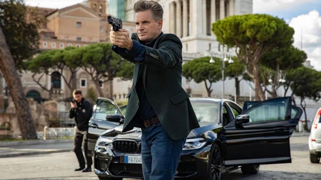Shea Whigham zielt in einer Stadt mit einer Pistole nach vorne, im Hintergrund ein schwarzes Auto und Bäume.