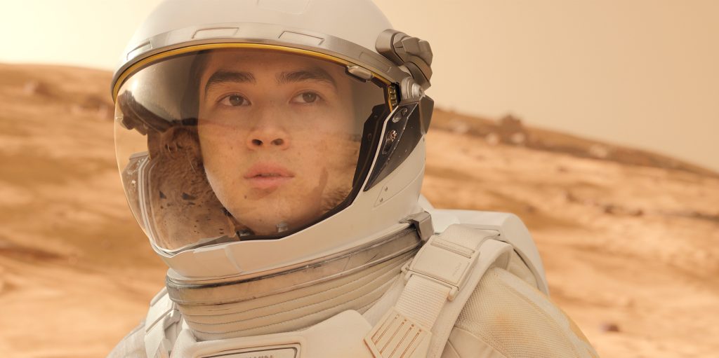 Nahaufnahme eines Astronauten in einem weißen Raumanzug mit Helm, der auf einer roten, sandigen Planetenoberfläche steht, möglicherweise dem Mars.