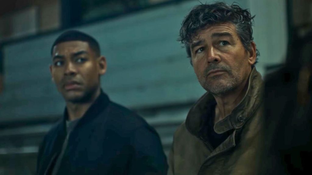 Zwei Männer blicken mit ernstem, konzentriertem Gesichtsausdruck nach rechts in einer schwach beleuchteten Umgebung.