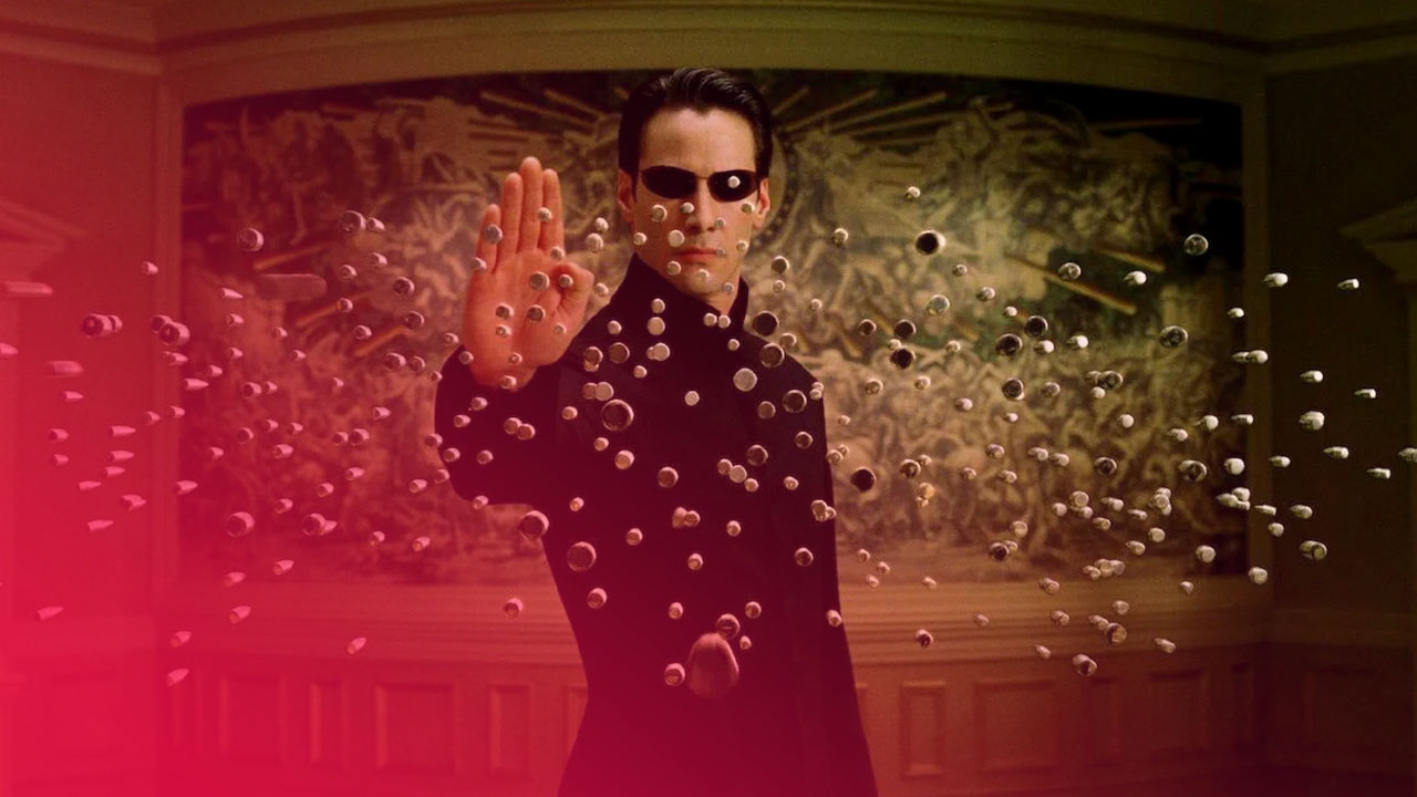 Ein Mann in schwarzem Mantel stoppt mit erhobener Hand Dutzende in der Luft schwebende Patronenkugeln vor einer Wand.