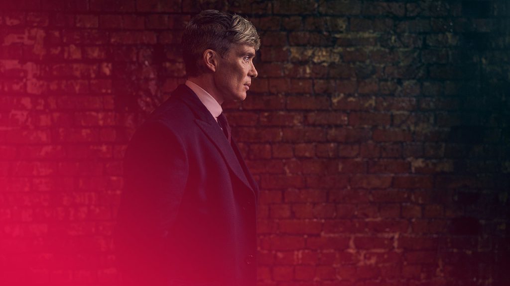 Ein Mann im dunklen Mantel, Hemd und Krawatte steht vor einer roten Backsteinmauer. Er blickt nach rechts. Das Bild hat dunkle Töne.