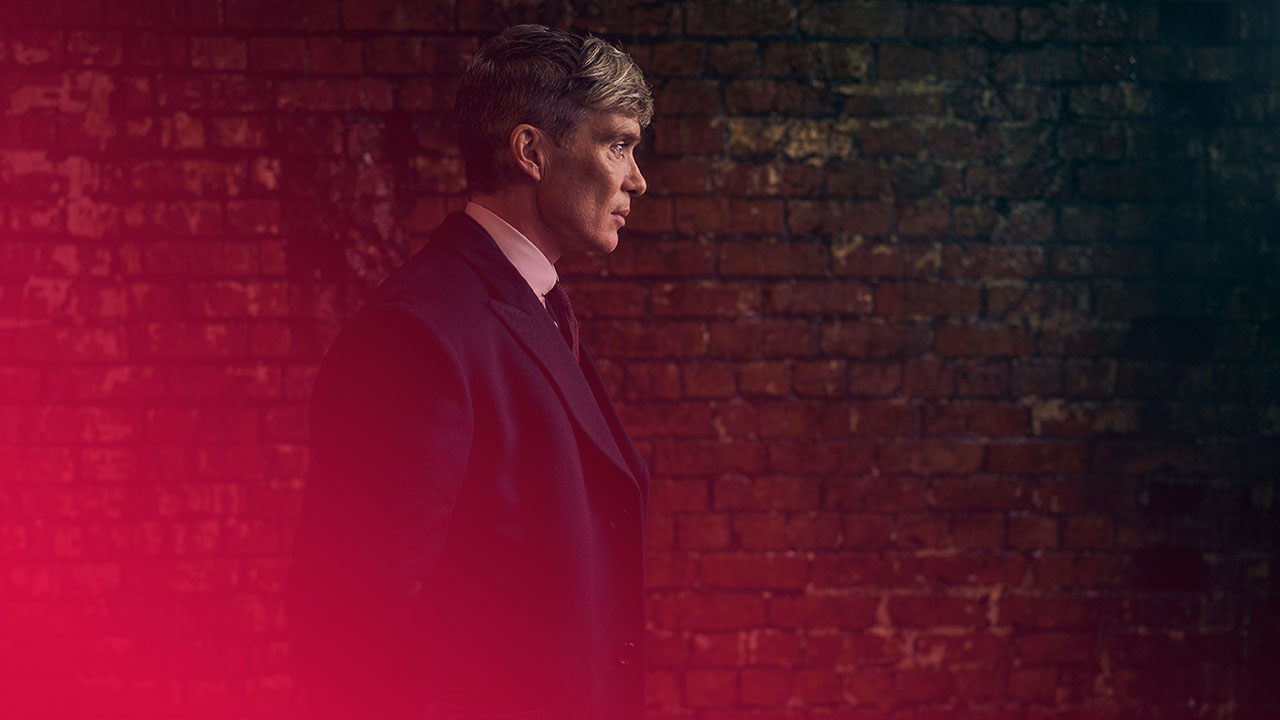 Ein Mann im dunklen Mantel, Hemd und Krawatte steht vor einer roten Backsteinmauer. Er blickt nach rechts. Das Bild hat dunkle Töne.