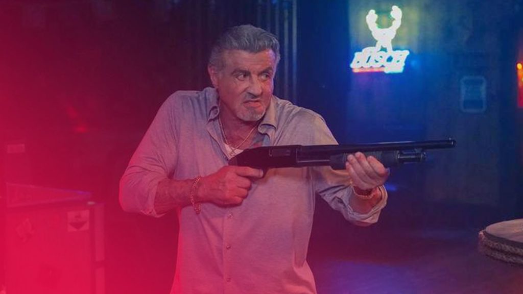 Älterer Mann mit grauem Haar und hellblauem Hemd zielt mit einer Schrotflinte. Im Hintergrund leuchtet ein blaues Neon-Schild.