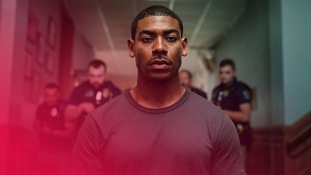 Ein dramatisches, mittig aufgenommenes Porträt eines Mannes mit hellem Teint in einem einfachen, dunkelgrauen T-Shirt, der direkt in die Kamera blickt. Er hat kurz geschnittenes Haar und einen ernsthaften Ausdruck. Im unscharfen Hintergrund stehen vier Männer in dunkelblauen oder schwarzen Uniformen, die Polizisten ähneln, mit ernsten Gesichtern. Die Szene scheint in einem Flur oder einem Innenraum mit hoher Decke und hellem Licht im Hintergrund aufgenommen worden zu sein, was auf eine spannungsgeladene oder konfrontative Situation hindeutet.