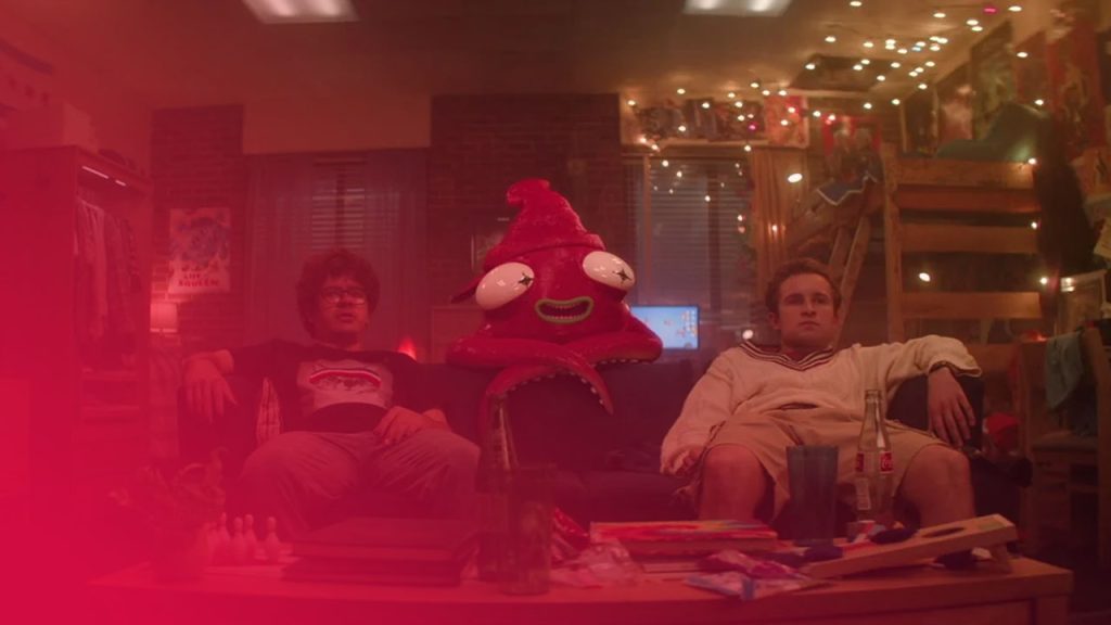 Eine große, rosa Tintenfisch-Figur mit großen Augen sitzt auf einem Sofa zwischen zwei jungen Männern in einem unordentlichen, warm beleuchteten Zimmer. Auf dem Couchtisch davor stehen Getränkeflaschen und verschiedene Gegenstände.