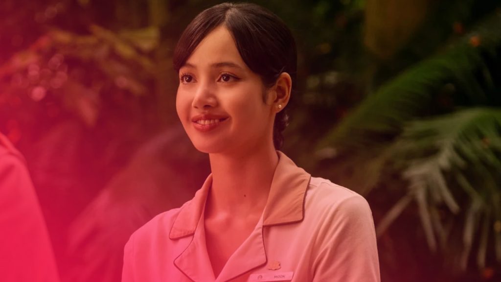 Lächelnde junge Frau in beiger Hoteluniform mit Namensschild vor tropischem, grünem Pflanzenhintergrund.