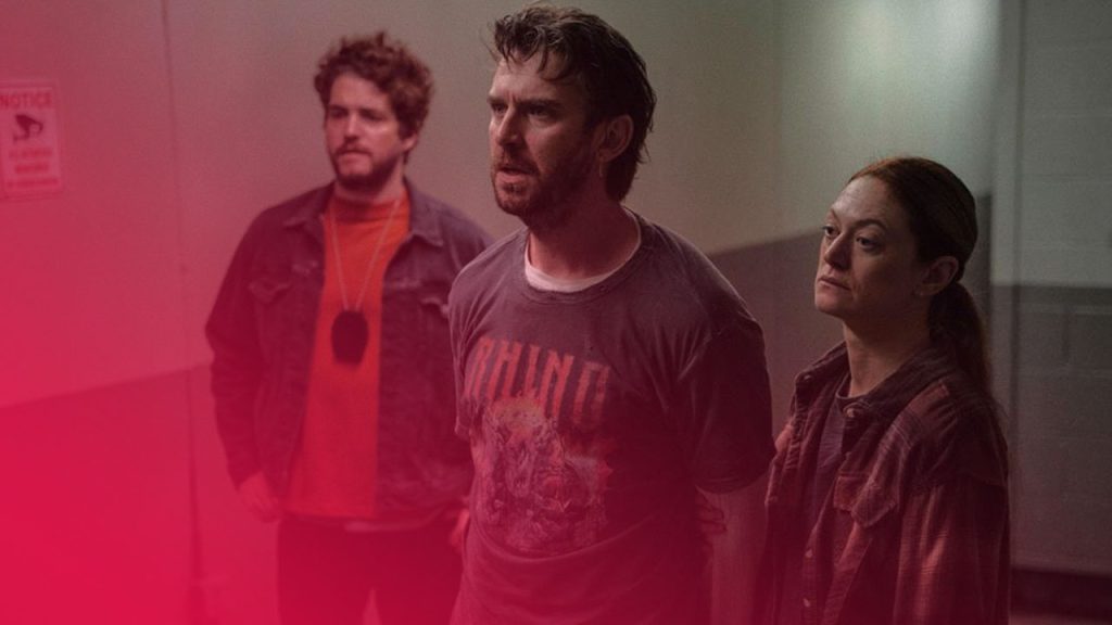 Drei Personen stehen in einem kahlen Flur: ein Mann mit zerrissenem T-Shirt im Vordergrund, eine Frau mit rotem Haar rechts neben ihm, und ein weiterer Mann im Hintergrund mit orangefarbenem Shirt und Jeansjacke, angespannte Stimmung.