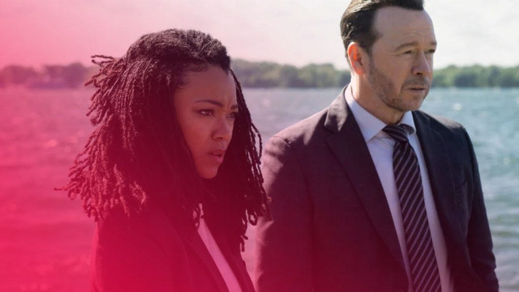 Eine Frau mit langen Dreadlocks und ein Mann im dunklen Anzug mit Krawatte stehen nebeneinander und blicken ernst in die Ferne, im Hintergrund ein sonnenbeschienener See mit einer Baumlinie am gegenüberliegenden Ufer.