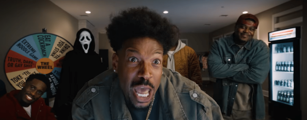 Vier Männer und eine Person in Ghostface-Maske vor einem Glücksrad; der Mann im Vordergrund schaut erschrocken.