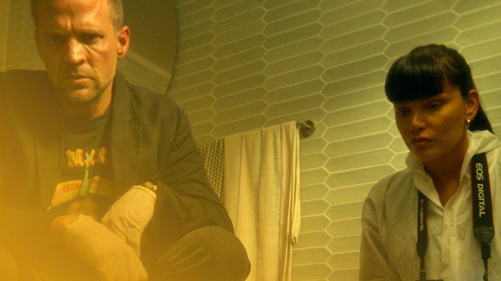 Ein Mann und eine Frau hocken in einem Badezimmer mit sechseckigen Fliesen. Der Mann trägt Handschuhe und blaue Überschuhe, die Frau einen weißen Schutzanzug und hält eine Kamera mit Blitz. Beide blicken konzentriert nach unten. Die Beleuchtung ist gelblich-grün.