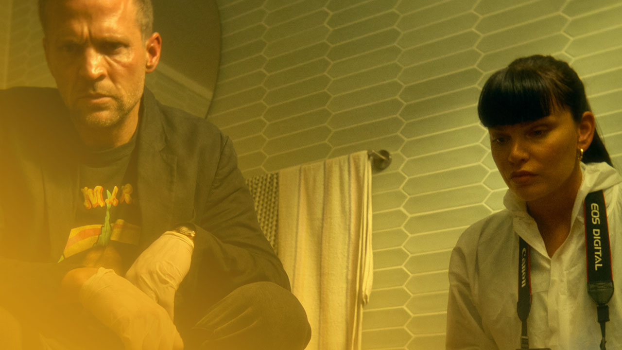 Ein Mann und eine Frau hocken in einem Badezimmer mit sechseckigen Fliesen. Der Mann trägt Handschuhe und blaue Überschuhe, die Frau einen weißen Schutzanzug und hält eine Kamera mit Blitz. Beide blicken konzentriert nach unten. Die Beleuchtung ist gelblich-grün.