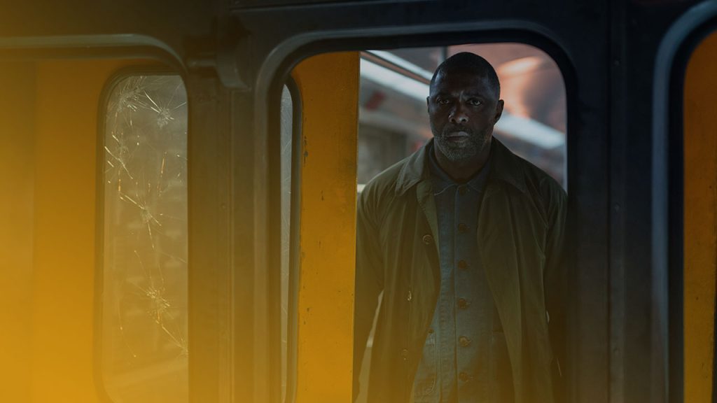 Mann im dunklen Mantel blickt ernst durch die Tür eines gelben Zugs mit gesplitterten Fensterscheiben.