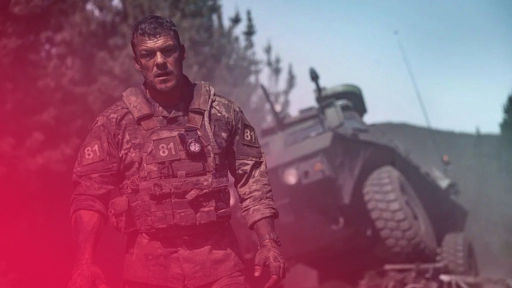 Alan Ritchson in Kampfmontur geht entschlossen vor einem umstürzenden Militärfahrzeug in rauchiger Umgebung.