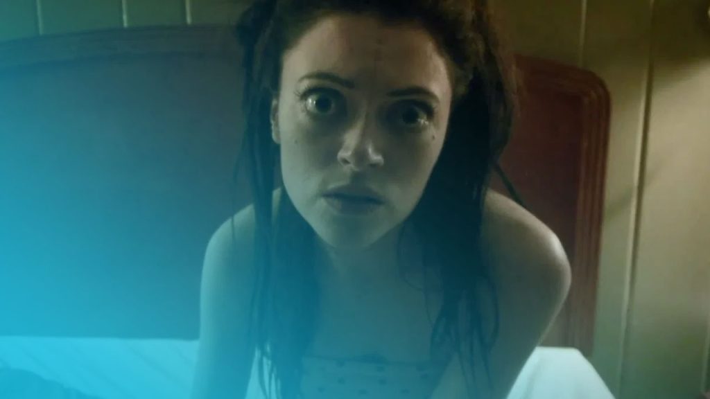 Frau mit dunklen, zerzausten Haaren und weit aufgerissenen Augen starrt erschrocken in die Kamera, sitzt aufrecht in einem Bett mit weißer Bettwäsche und einem hölzernen Kopfteil im Hintergrund.