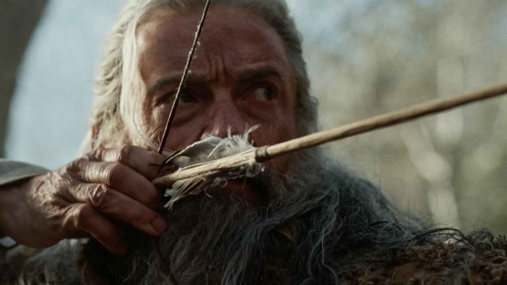 Nahaufnahme eines älteren Mannes mit langem grauem Bart, der konzentriert einen Bogen mit aufgelegtem Pfeil spannt.