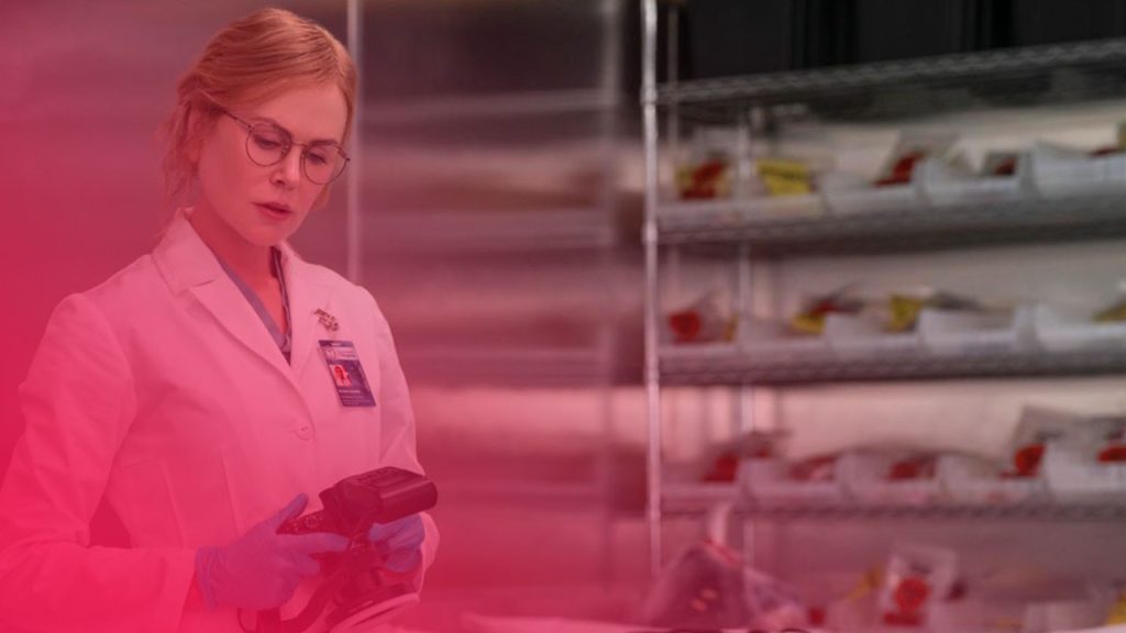 Frau in weißem Kittel, Brille und blauen Handschuhen hält eine Kamera. Eine Leiche ist mit einem weißen Tuch bedeckt. Hinter ihr ein Regal mit Behältern.